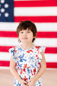 Toddler girl in Born to Sparkle Flutter Sleeve Twirl Dress, patriotic print, posed before an American flag backdrop.