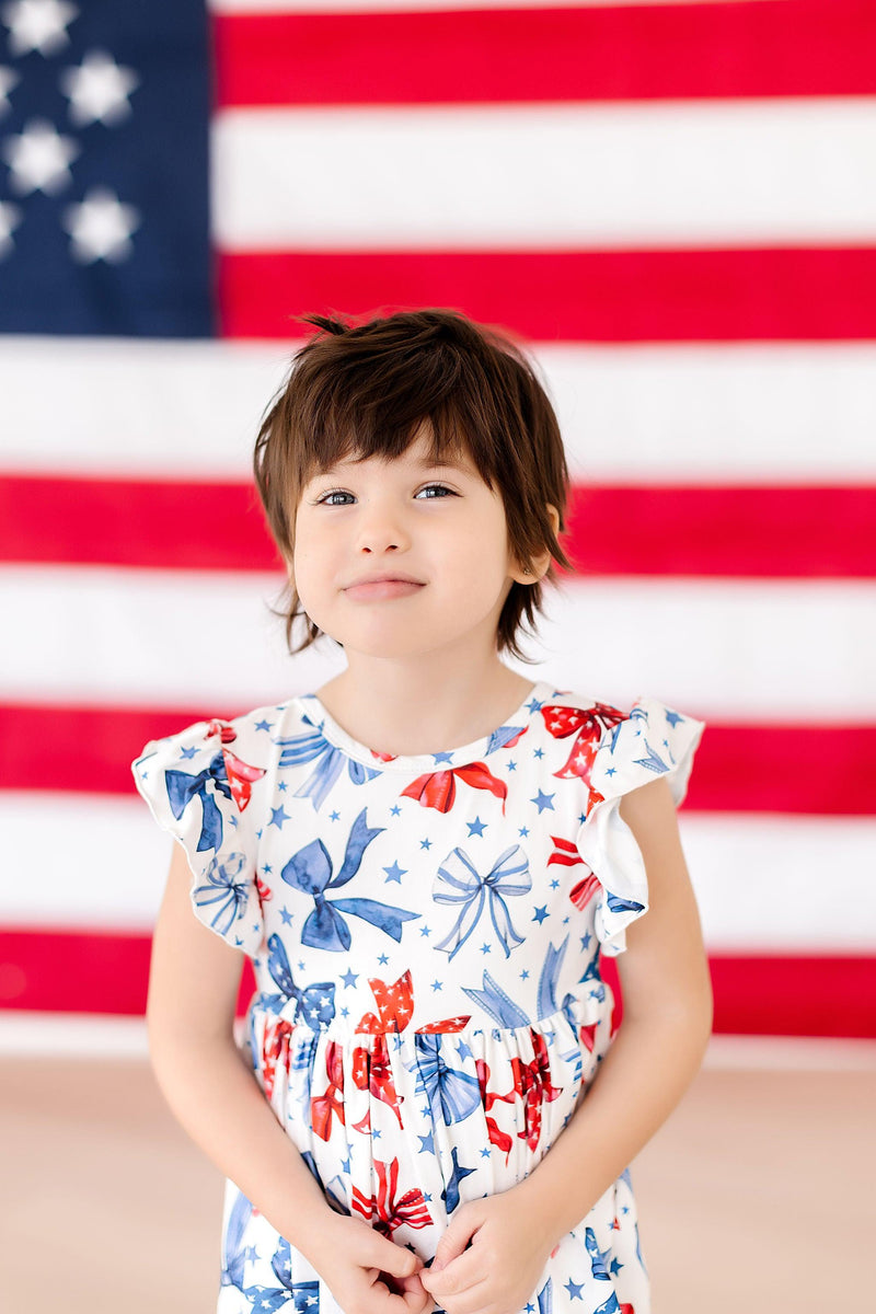 Toddler girl in Born to Sparkle Flutter Sleeve Twirl Dress, patriotic print, posed before an American flag backdrop.