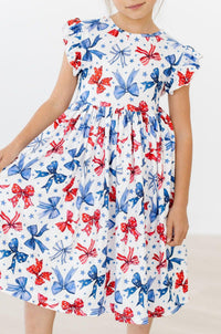 Born to Sparkle Flutter Sleeve Twirl Dress worn by a toddler girl; white fabric with red and blue bows and stars, twirly.