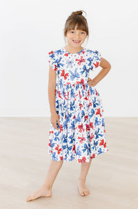 Smiling girl in a white Born to Sparkle Flutter Sleeve Twirl Dress with red and blue bow pattern.