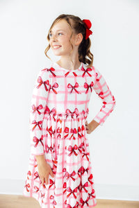 Girl wearing Bow La La Long Sleeve Pocket Twirl Dress with pink bows on white plaid.