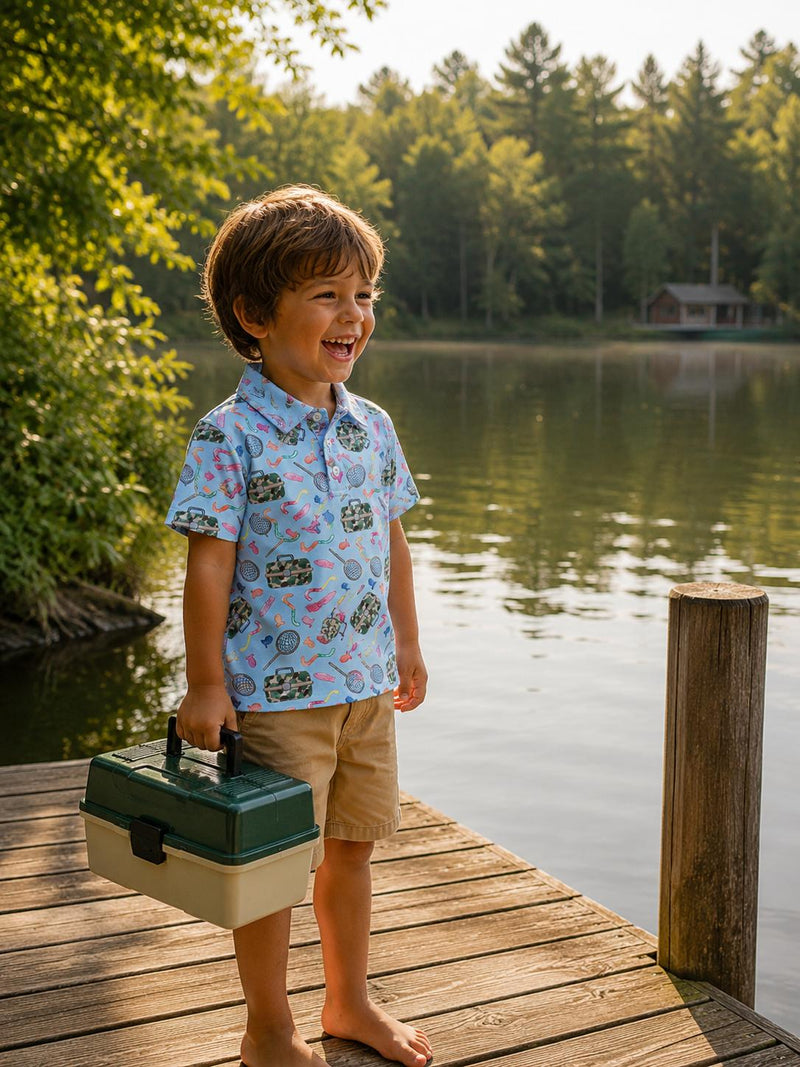 Boy in Boys Fishing Snacks S/S Polo stands on a lakeside dock, khaki shorts, holding a tackle box.