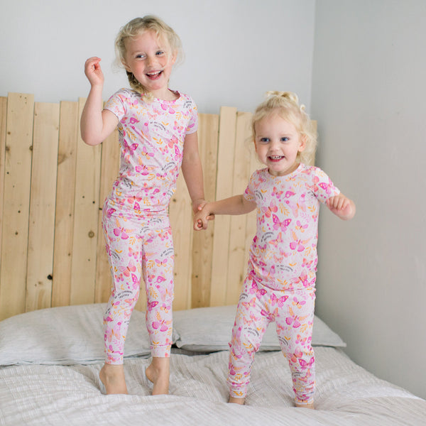 Two children in pink butterfly-print pajamas from The Second-Skin 2-Piece Short Sleeve Pajama set, smiling on a bed.
