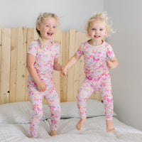 Two children wear The Second-Skin 2-Piece Short Sleeve Pajama | Butterfly Smoochies in pink butterfly print, indoors on a bed.