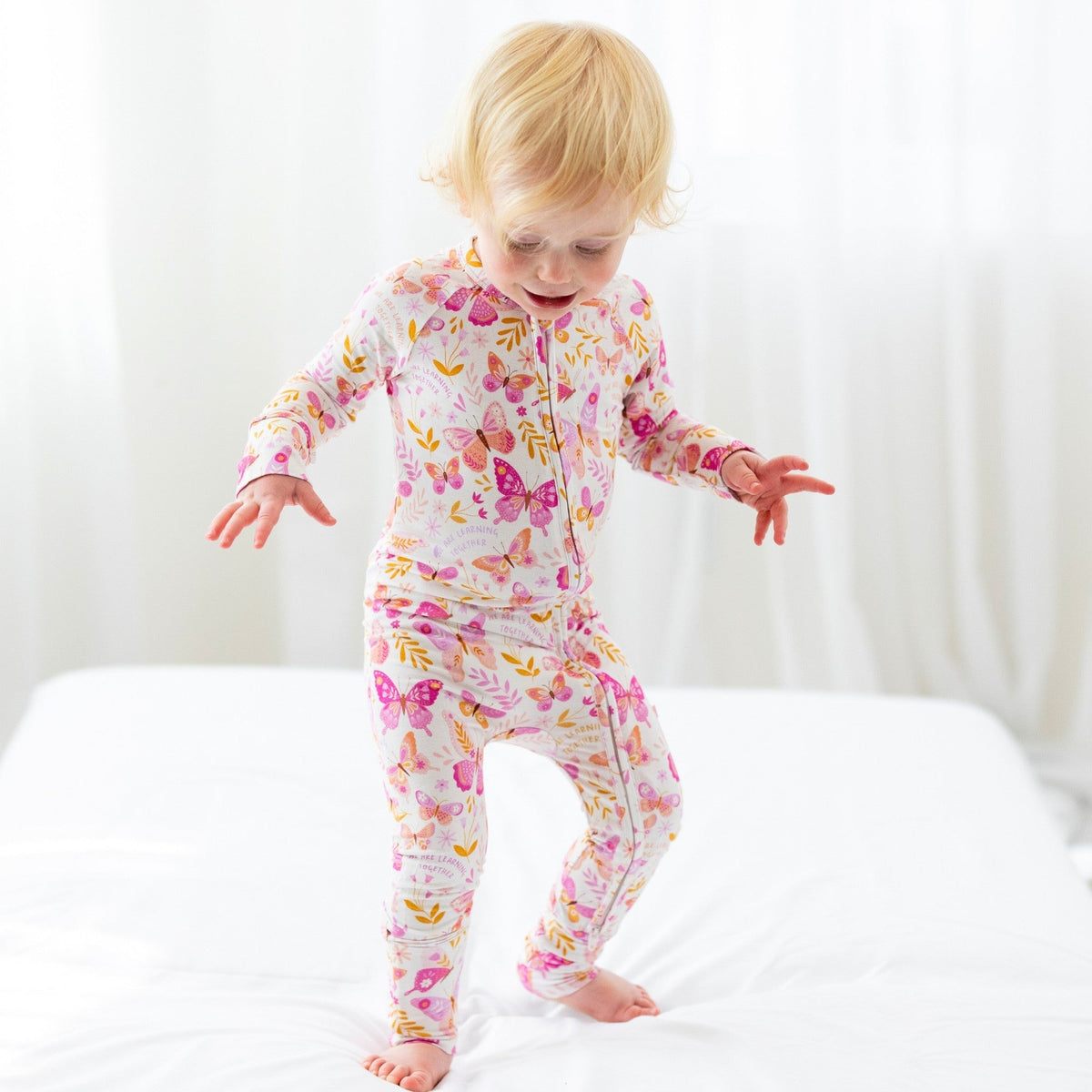 The Second-Skin Convertible Romper butterfly print with zipper, worn by a toddler on a white bed.