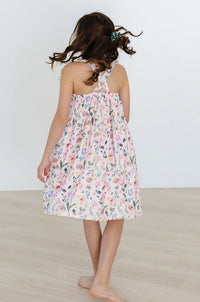 Girl wearing Butterfly Meadows Ruffle Cross Back Dress with crisscross straps and floral print.