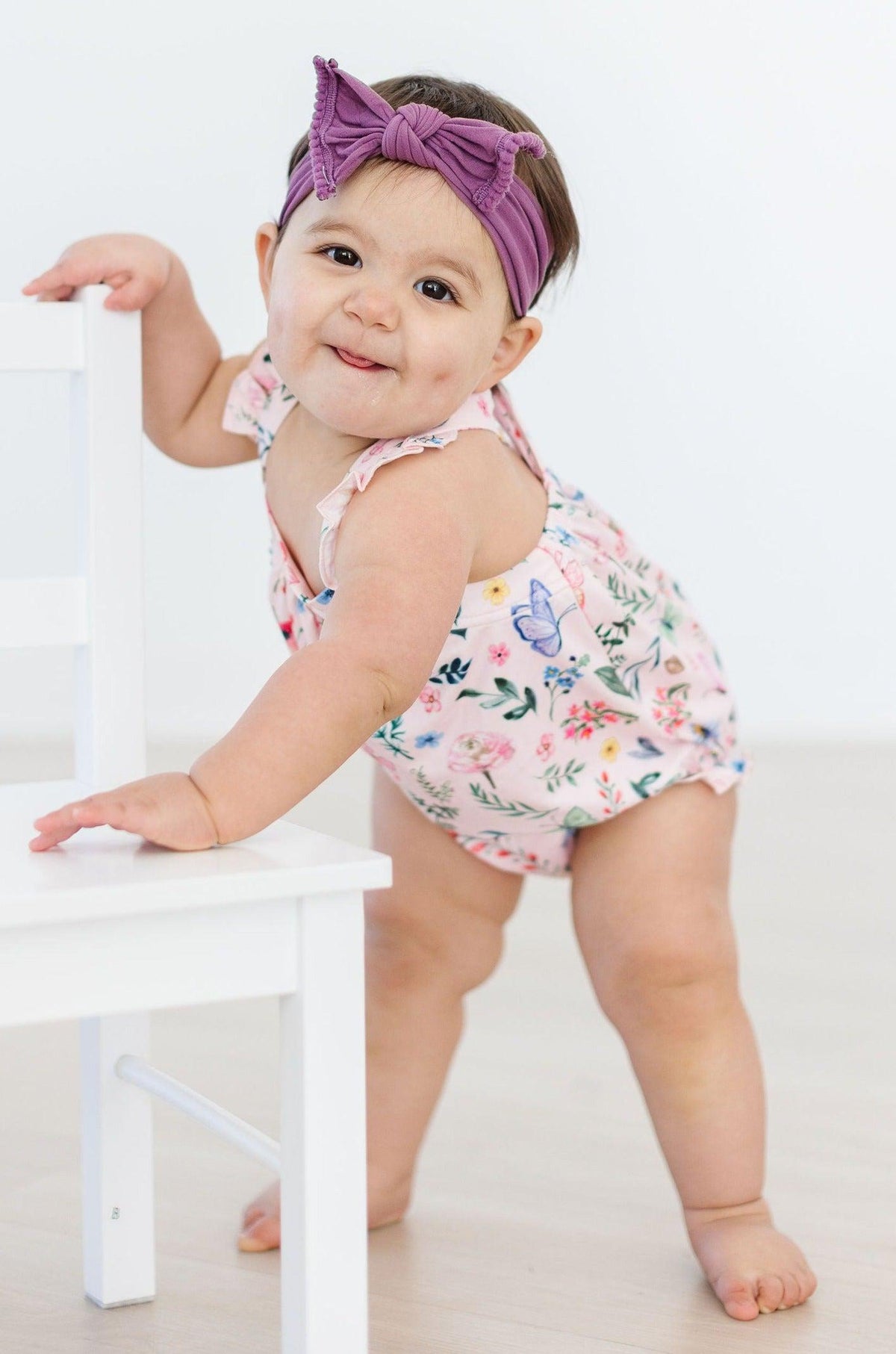 Butterfly Meadows Ruffle Cross Back Romper on a baby girl in a floral romper with a purple headband.