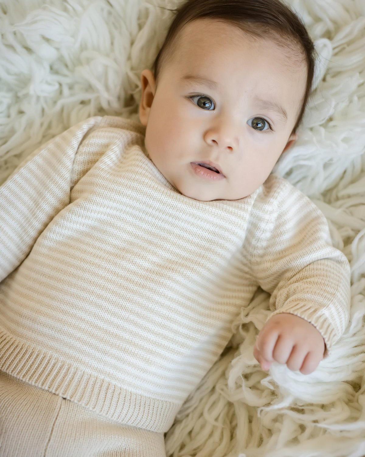 Baby wearing oatmeal and cream stripe knit sweater with raglan sleeves and solid oatmeal pull-on pants, resting on a soft white textured rug.