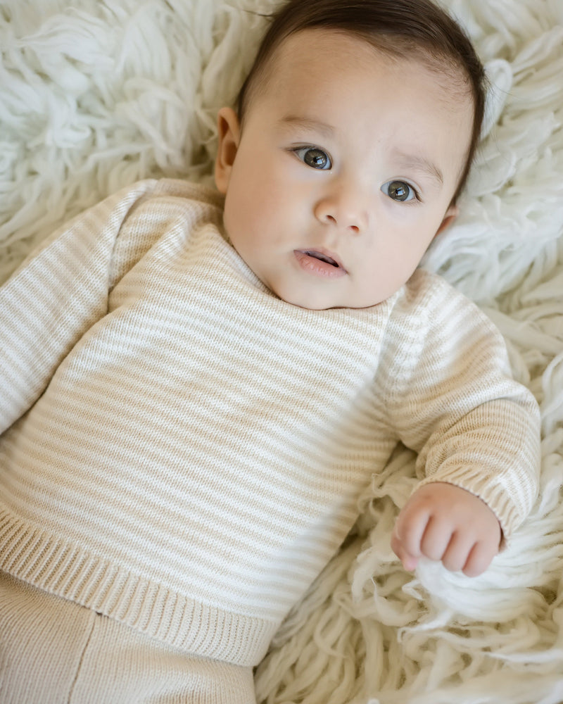 Baby wearing oatmeal and cream stripe knit sweater with raglan sleeves and solid oatmeal pull-on pants, resting on a soft white textured rug.