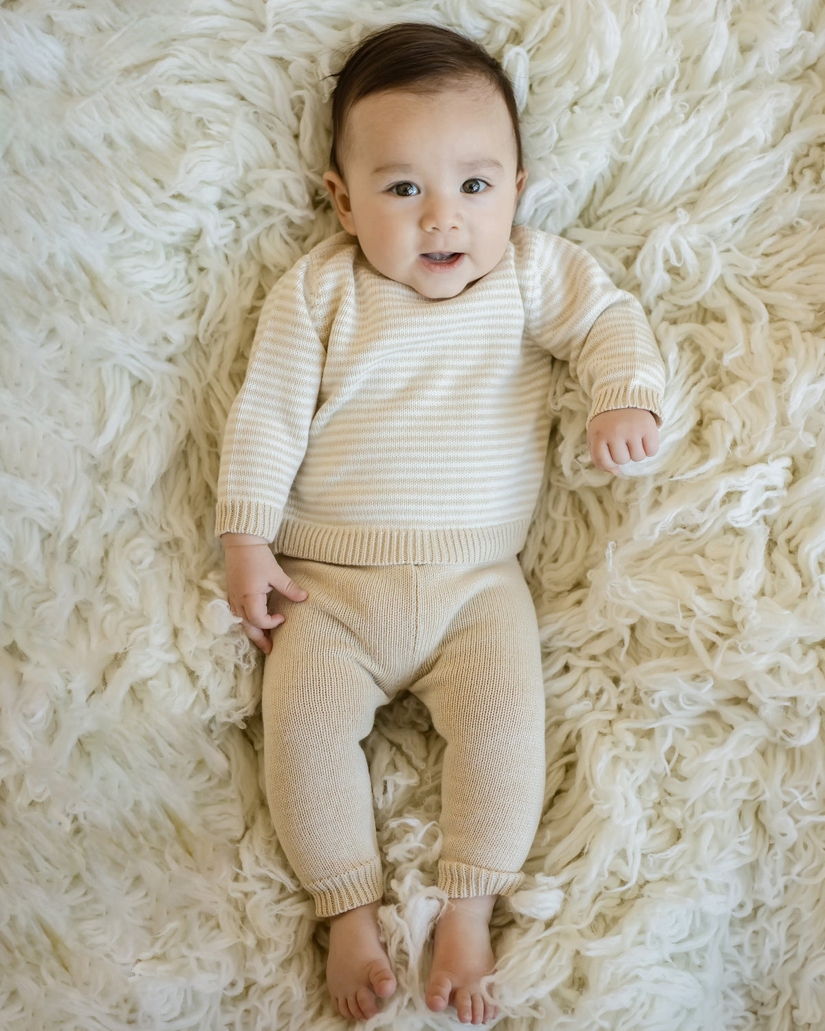 Baby wearing oatmeal stripe knit sweater and solid oatmeal pull-on pant set lying on fluffy white rug.