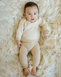 Baby wearing oatmeal stripe knit sweater and solid oatmeal pull-on pant set lying on fluffy white rug.