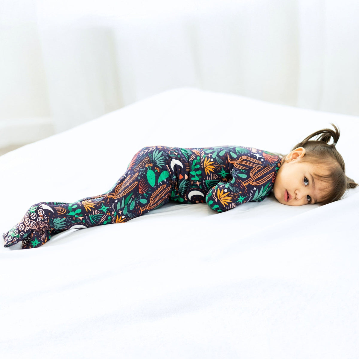 Child wearing Desert Cactus Midnight romper with tropical print on a white bed.