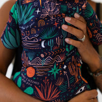The Second-Skin Footie Desert Cactus Midnight worn by mom; zippered one-piece with "Mama, I Wished for You" print.