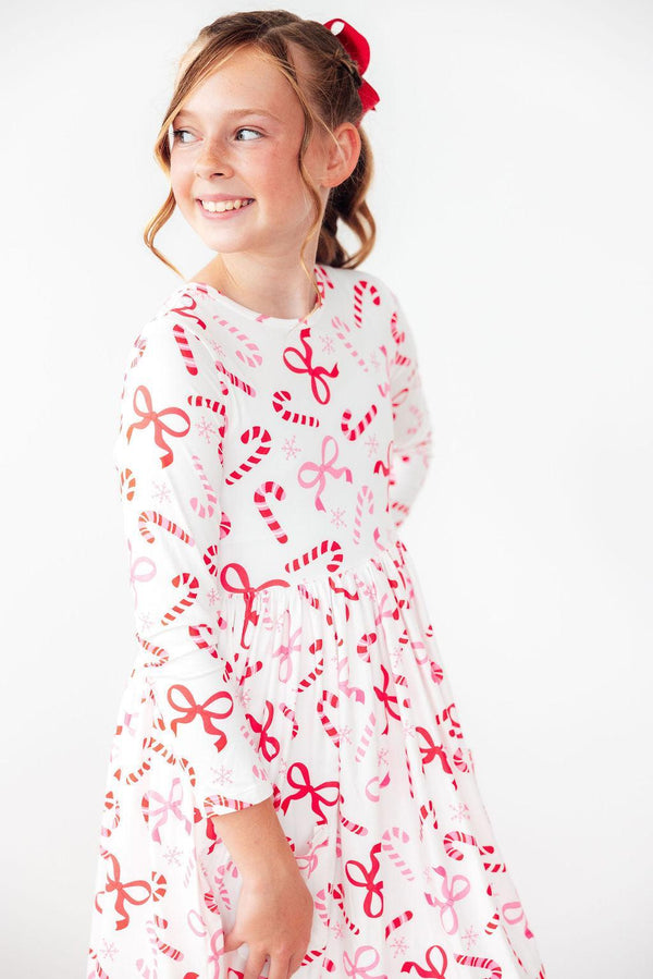 Girl wearing white long sleeve twirl dress with red candy cane, bow, and snowflake print. Features soft stretch fabric and full circle skirt for festive comfort.