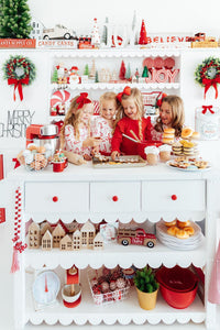 Children in red and white holiday prints decorate cookies at a festive white baker’s play kitchen filled with Christmas decor.