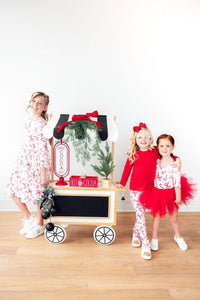 Girls modeling candy cane print long sleeve pocket twirl dresses and matching pajamas beside a festive hot cocoa cart.