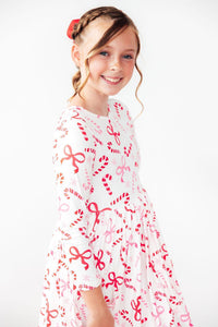 Girl smiling in white long sleeve twirl dress with red candy cane and bow print, featuring a full circle skirt and festive holiday design.