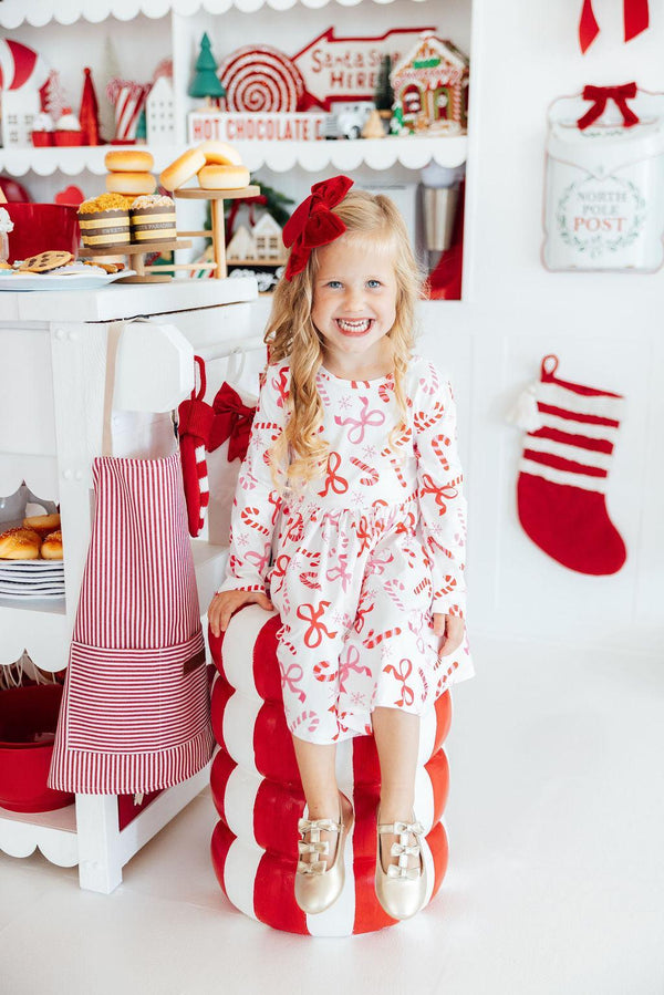 Girl in candy cane print long sleeve twirl dress with pocket, sitting on giant prop in festive candy shop setting.