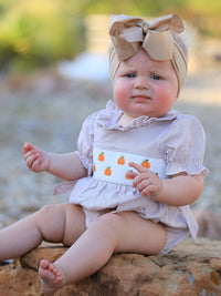Baby girl in plaid, fall pumpkin-embroidered smock romper with a beige bow headwrap sits on rocks outdoors.