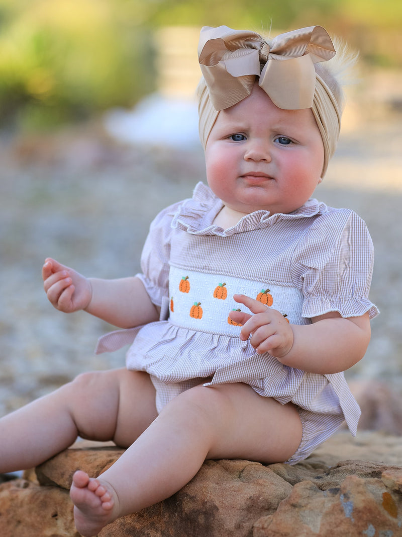 Baby girl in plaid, fall pumpkin-embroidered smock romper with a beige bow headwrap sits on rocks outdoors.