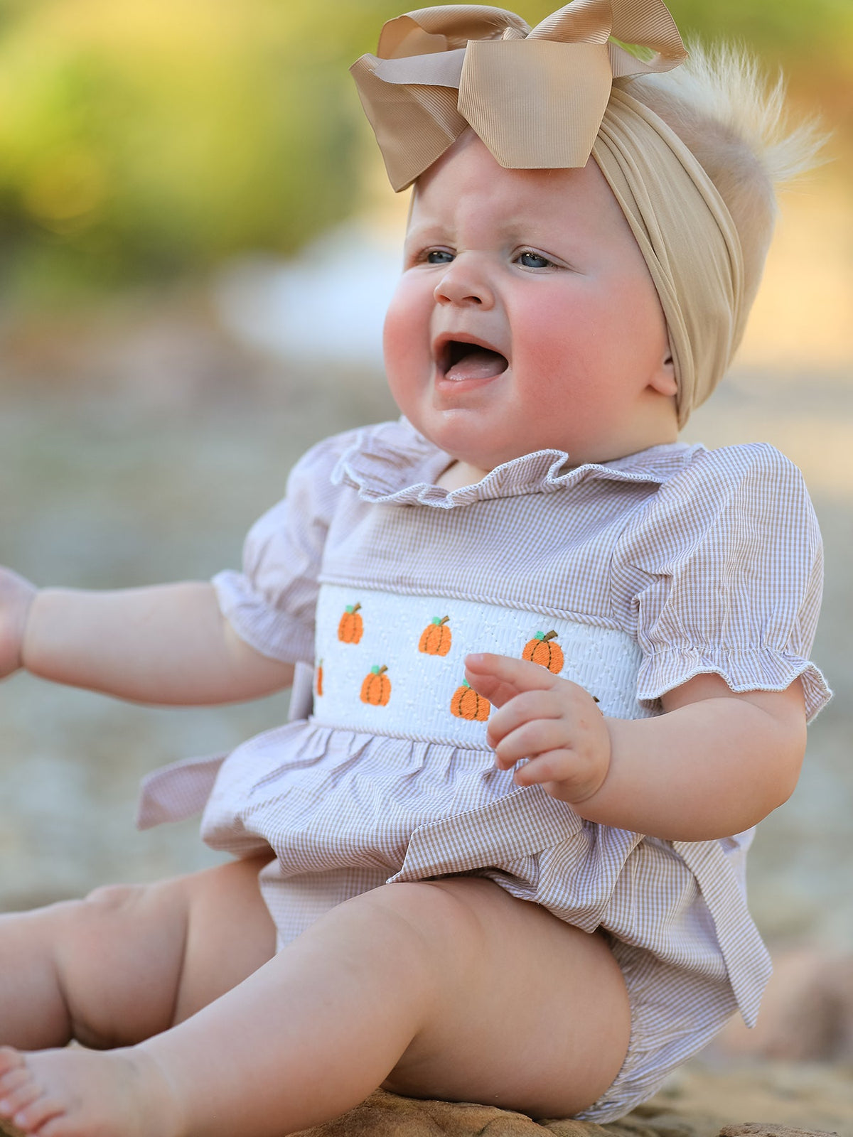 Baby girl in plaid pumpkin-embroidered smock romper with beige headband, ideal for fall Halloween and Thanksgiving.