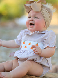 Baby girl in plaid pumpkin-embroidered smock romper with beige headband, ideal for fall Halloween and Thanksgiving.