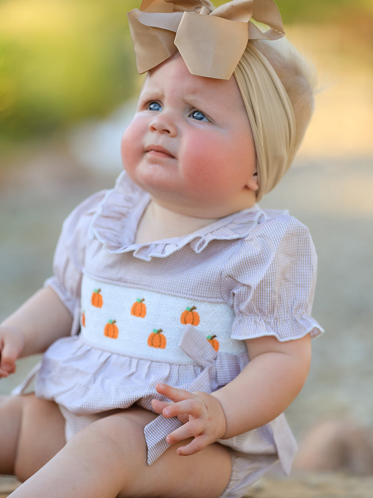 Baby girl in plaid smocked romper with pumpkin embroidery and a beige bow headband.
