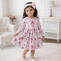 A young girl wearing a long-sleeve pink floral dress with a matching pink bow headband and light pink ballet flats, standing in a bright, white, classic-style room.