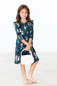 Girl wearing a dark green Christmas ballet dress with ruffle 3/4 sleeves and nutcracker print, holding a white handbag.