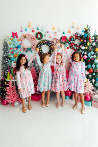 Four girls wearing Cocoa Kisses Long Sleeve Pocket Twirl Dress pose in front of colorful holiday wreaths and trees.