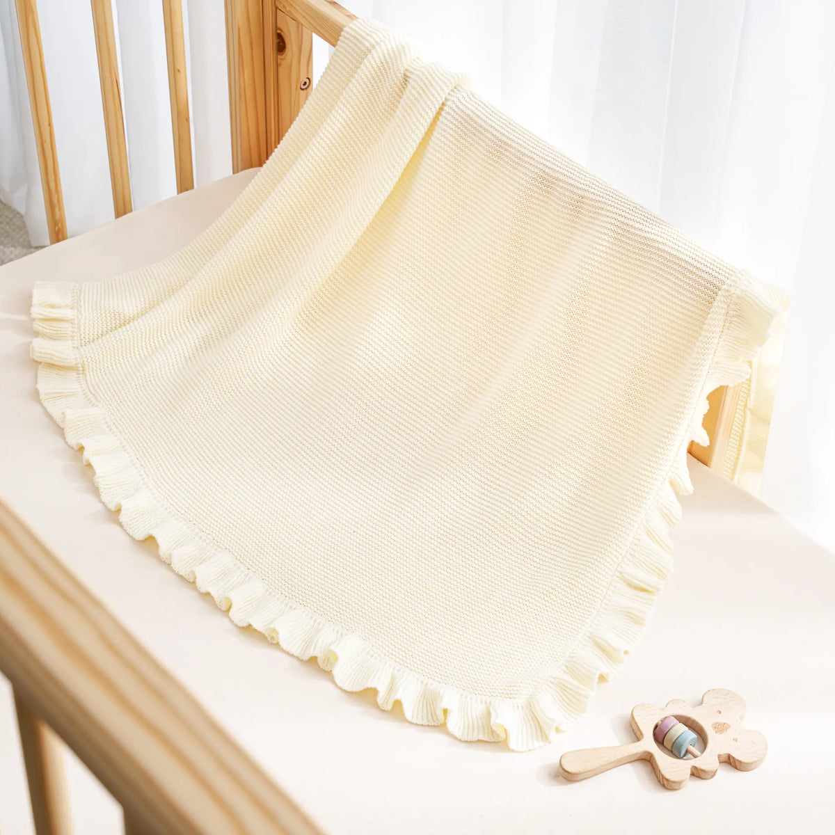 Cream ruffled edge baby blanket draped over light wooden crib rail with flower rattle
