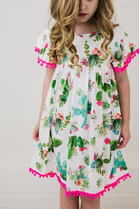 Cute Cactus Pom Pom Dress worn by a young girl, white fabric with cactus and floral print and hot pink pom-pom trim.