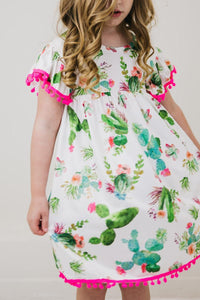 Cute Cactus Pom Pom Dress worn by a little girl, white dress with cactus print and pink pom pom trim.