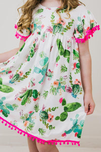 Little girl in Cute Cactus Pom Pom Dress featuring cactus and floral print with bright pink pom-pom trim.