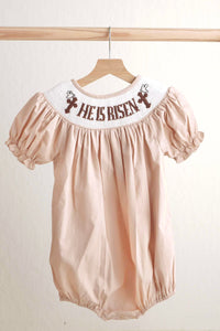 Pink baby romper with 'He Is Risen' text on a wooden hanger against a white background