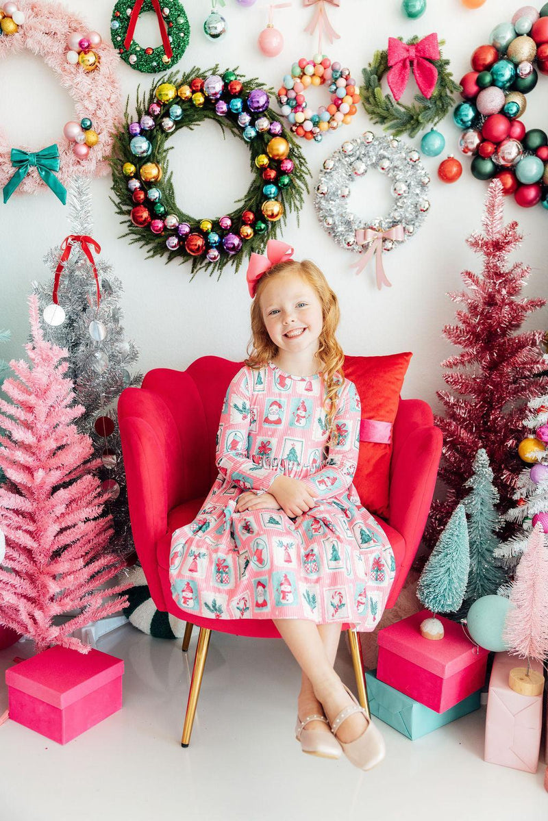 Smiling girl in pink long sleeve pocket twirl dress with holiday print, sitting in red chair surrounded by Christmas wreaths and trees.