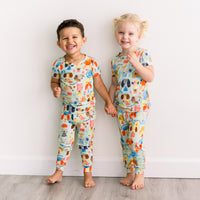 Two children in The Second-Skin 2-Piece Short Sleeve Pajama set by Mindful Doggos, colorful hand-drawn animal print.