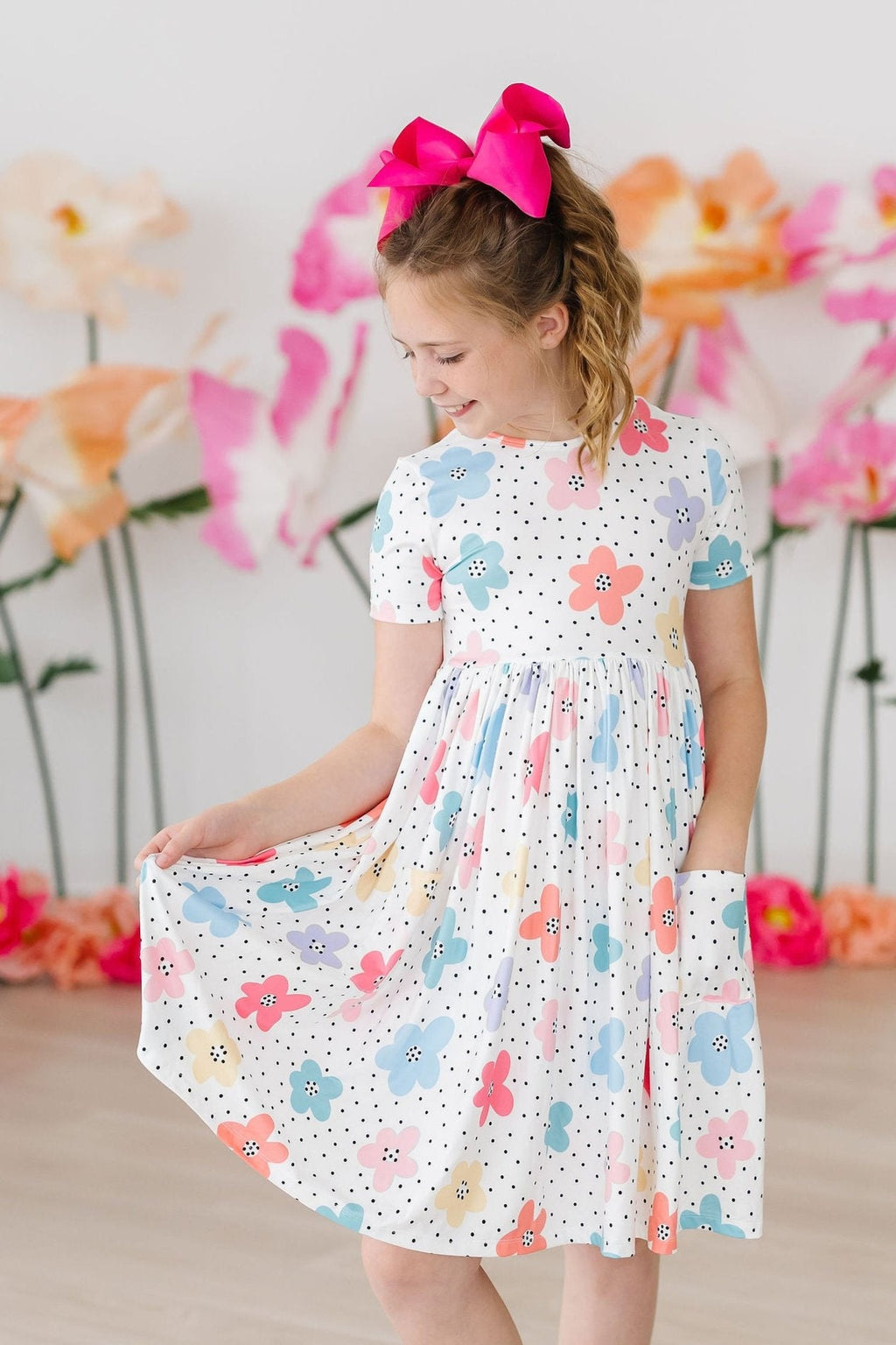 A cheerful girl wearing the Dots & Daisies S/S Pocket Twirl Dress by Mila & Rose, showcasing a playful mix of colors and patterns. The dress features a fun floral design and a handy pocket, complemented by a stylish pink bow in her hair.