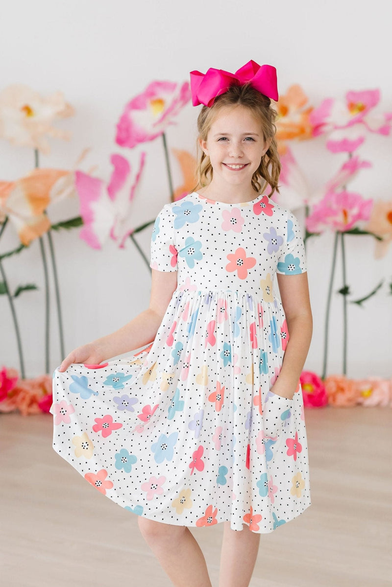A cheerful girl posing in the Dots & Daisies S/S Pocket Twirl Dress, featuring vibrant flower patterns, complete with a playful pocket and a bright pink bow.