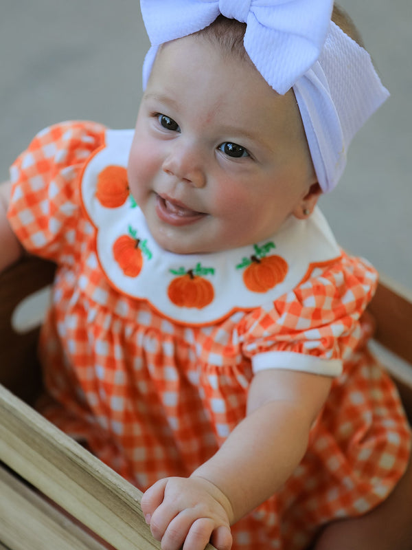 Baby girl wearing orange gingham bloomers with puff sleeves, scalloped Peter Pan collar, and pumpkin applique, paired with a white bow headband.