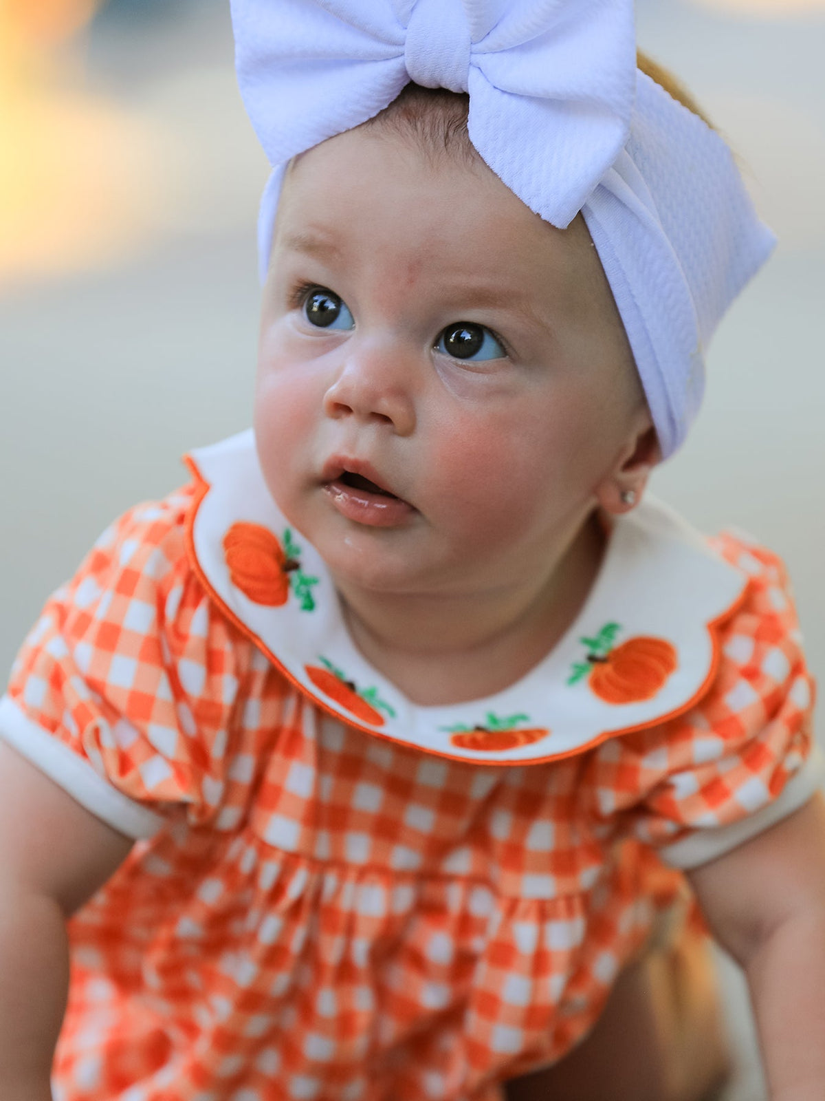 Baby girl in orange gingham puff sleeve outfit with pumpkin applique Peter Pan collar and lavender bow headband.
