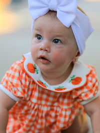 Baby girl in orange gingham puff sleeve outfit with pumpkin applique Peter Pan collar and lavender bow headband.