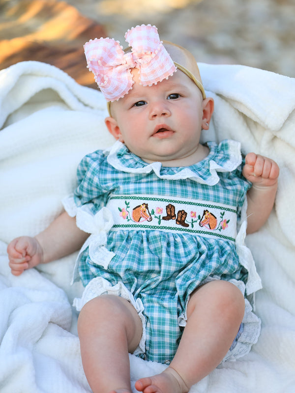 Baby girl wearing mint plaid bubble romper with horse and cowboy boot embroidery, lace trim, and pink gingham bow headband.