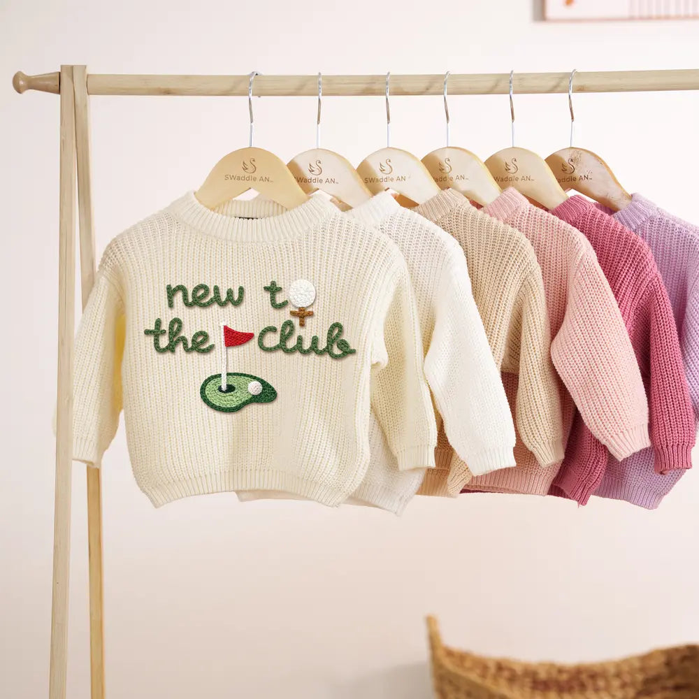 Cream sweater on wooden hanger with green 'new to the club' embroidery, red flag, and ball tee next to beige and pink