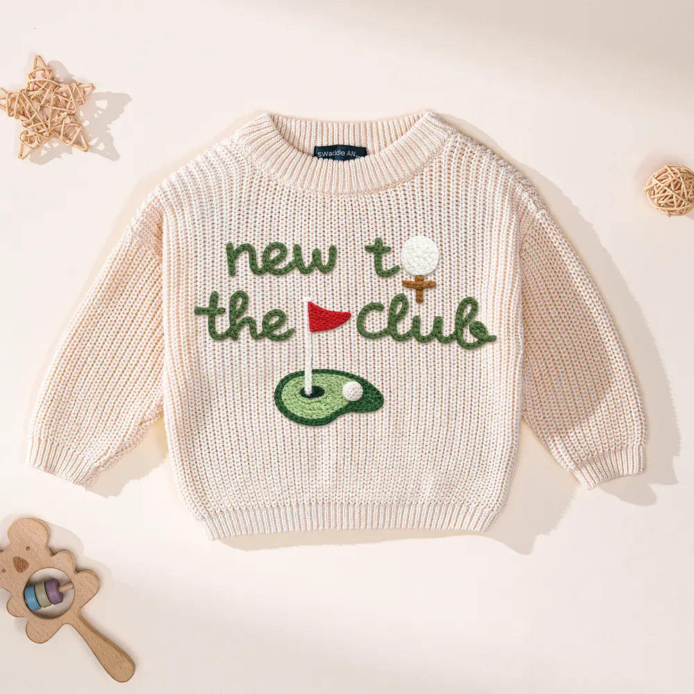 Top-down view of oatmeal knit sweater with green lettering and colorful golf embroidery beside rattan balls and wooden toy