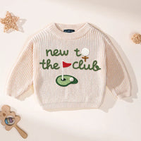 Top-down view of oatmeal knit sweater with green lettering and colorful golf embroidery beside rattan balls and wooden toy