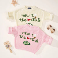 Flat-lay of overlapping cream and pink sweaters showcasing green embroidered 'new to the club' text and golf graphics with