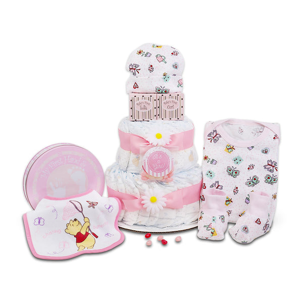 A baby gift set arranged on a white background, featuring a diaper cake made of folded diapers with pink ribbons and a Winnie the Pooh flower topper, a pink round tin, a printed bib with Winnie the Pooh, and a pink patterned baby onesie.