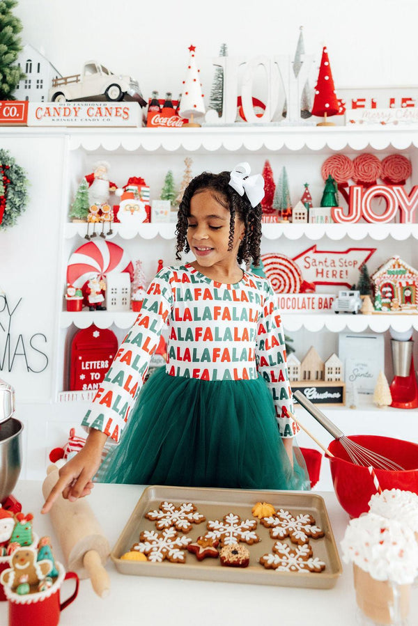 Little girl in "Fa La La" long sleeve dress with green tulle skirt baking holiday cookies in a festive kitchen.
