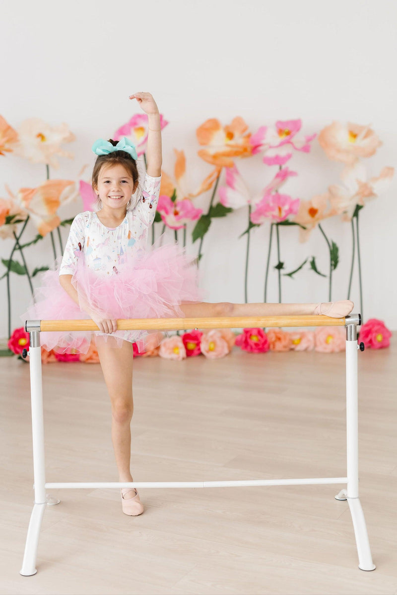 A joyful girl in a Fairy Princess 3/4 Sleeve Tutu Leotard by Mila & Rose, performing a dance pose at a ballet barre, with vibrant flower decorations in the background. Perfect for twirls and magical moments!
